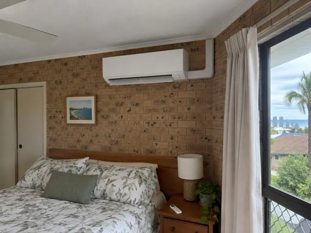 Wall-mounted split system above a bed in a brick bedroom with curtains and a window view in Burleigh Heads